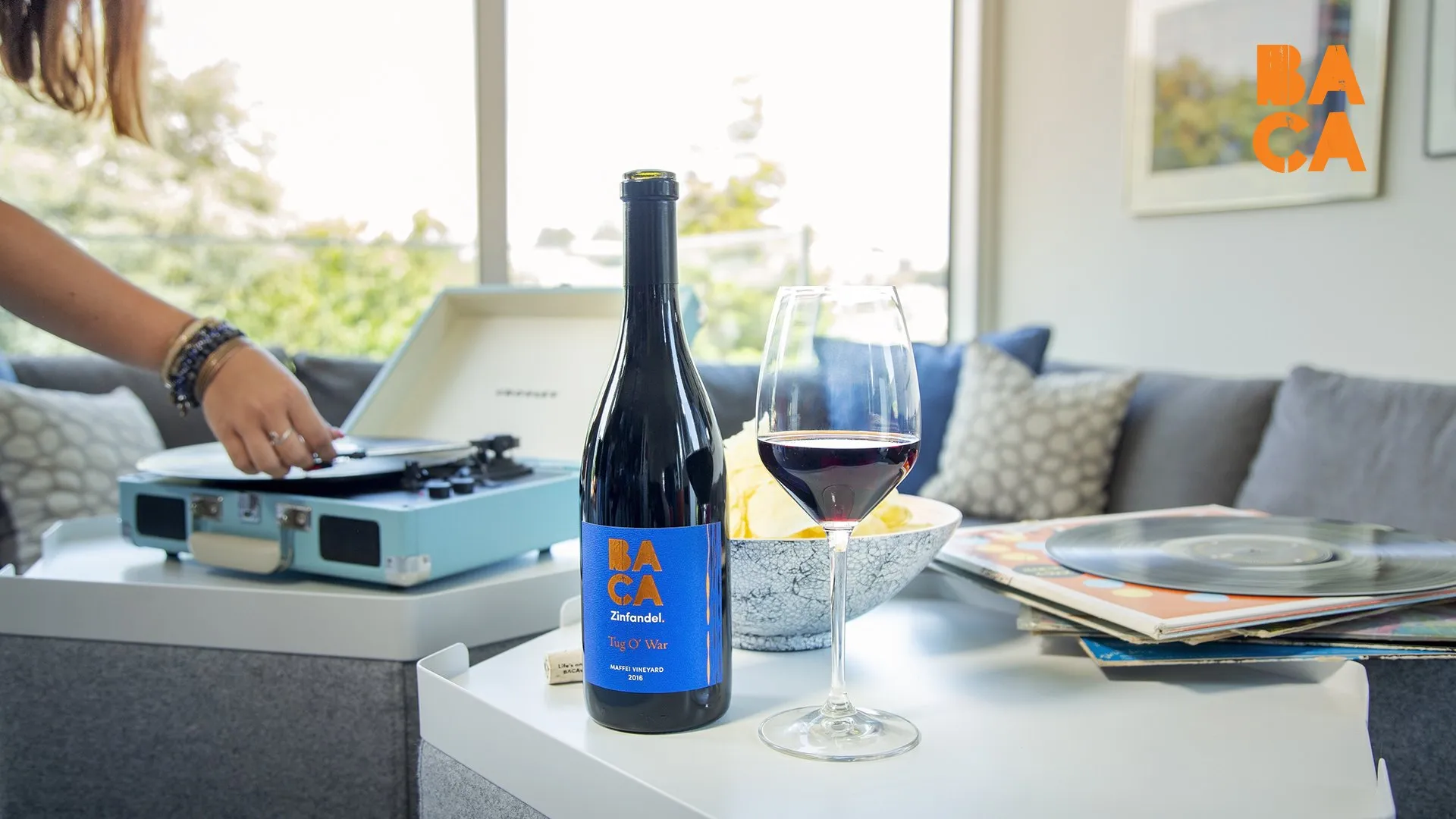 A bottle of BACA Tug O’ War Zinfandel and a glass of red beside a record player, a stack of vinyl on the table