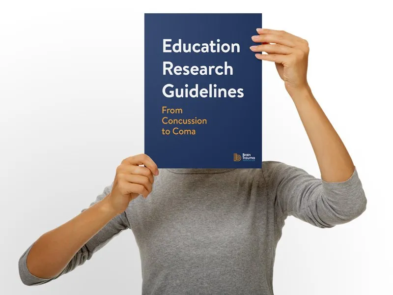 A person holding a navy booklet over their face: Education, Research, Guidelines, From Concussion to Coma