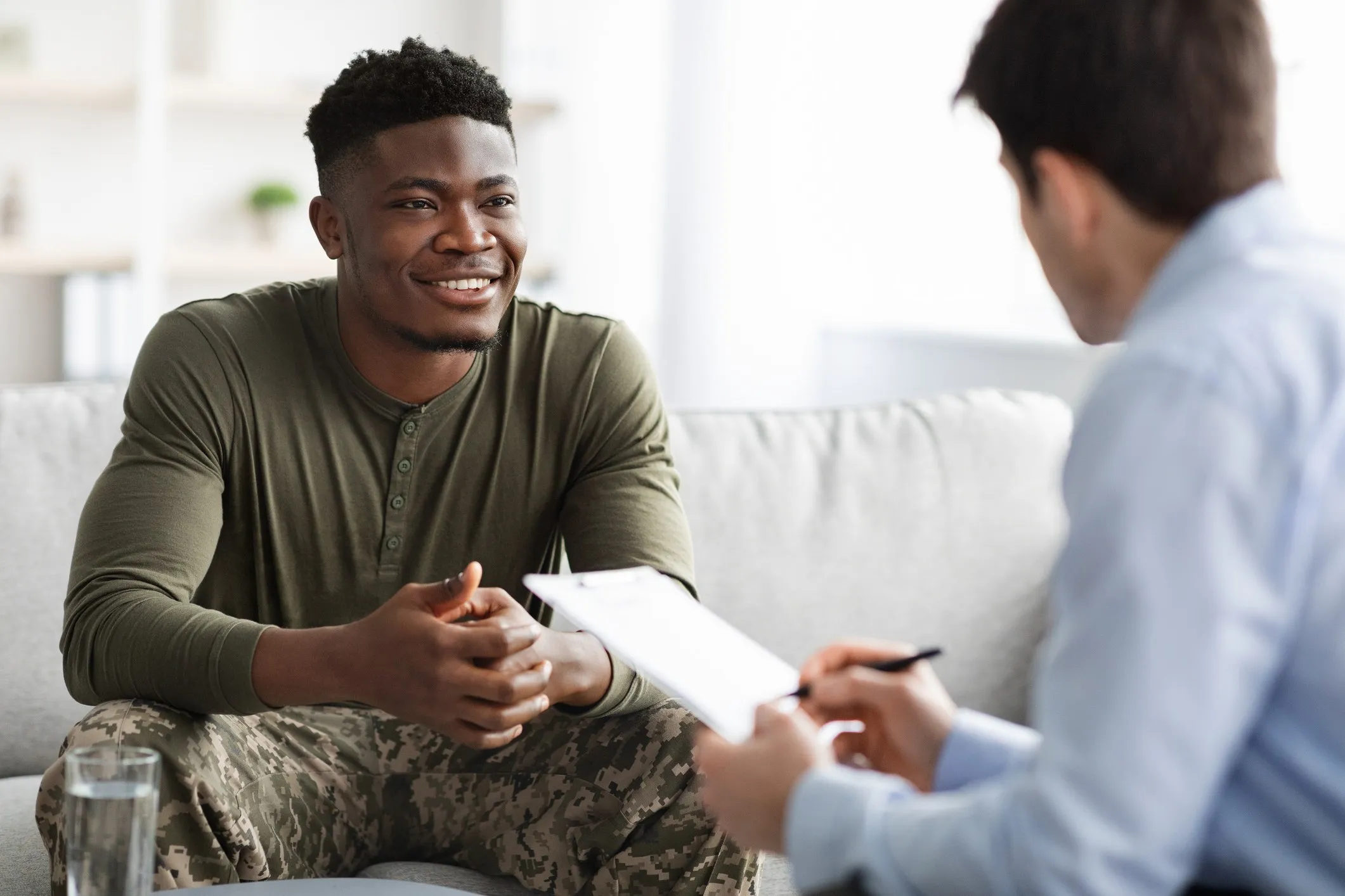 A smiling veteran in an olive shirt and camouflage pants talks with a counselor who takes notes