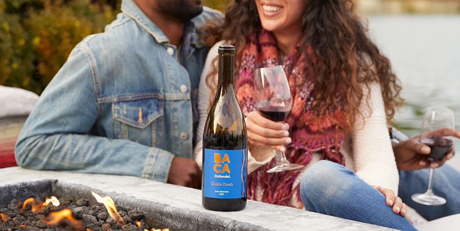 Two friends by a fire pit with a bottle of BACA Double Dutch and glasses of red