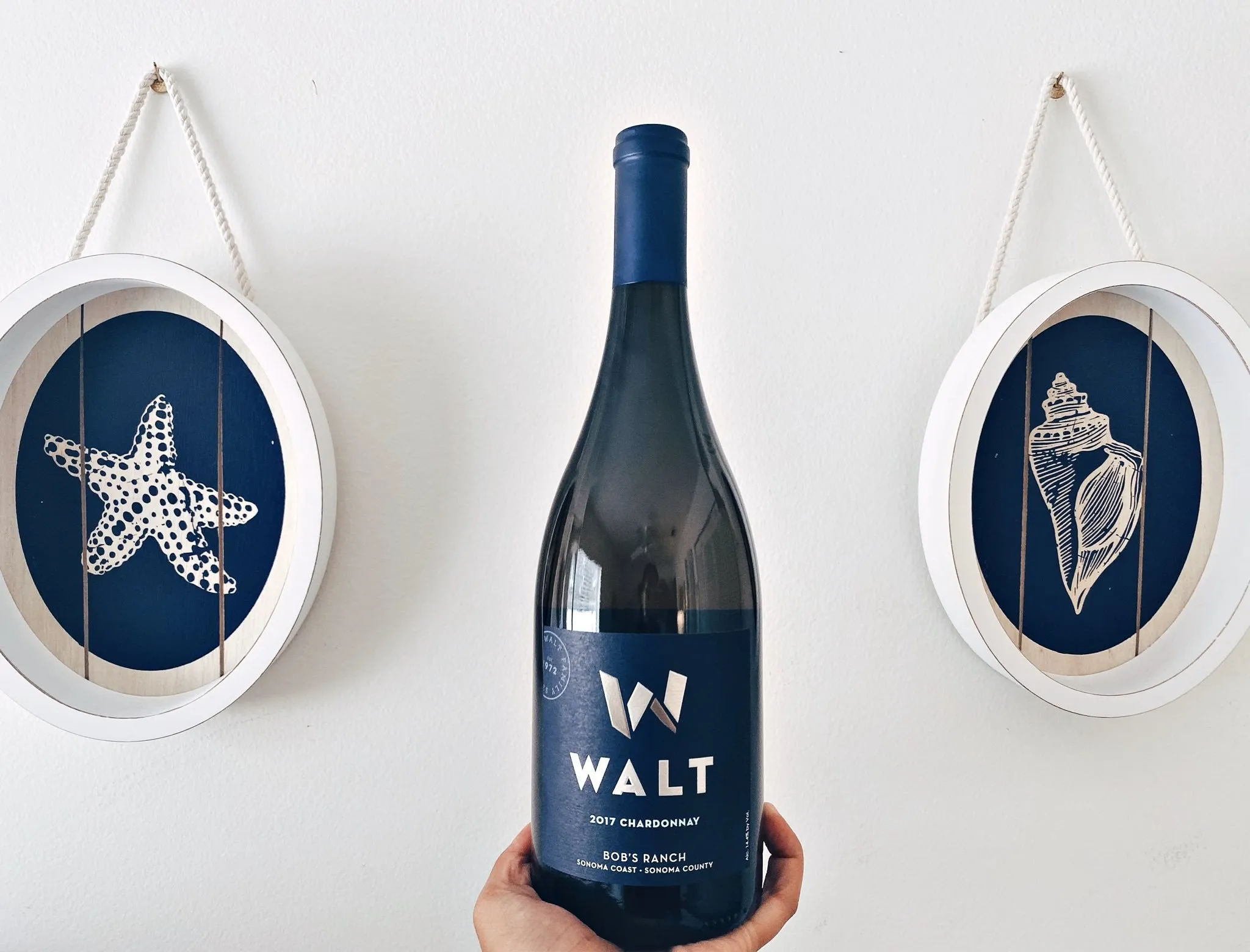 A hand holding a navy-labeled bottle of WALT Bob’s Ranch Chardonnay between two nautical prints